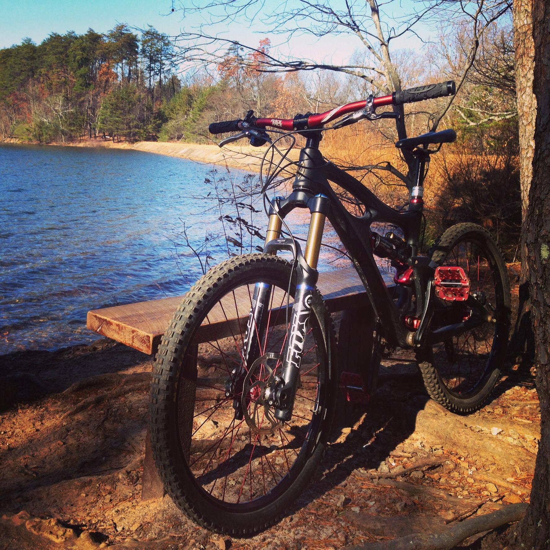 Ibis Mojo HD: A mountain bike with black and red accents is parked near a calm lake, surrounded by trees and grassy areas. The sun shines brightly, reflecting off the water, while a wooden bench is visible in the background, adding to the serene outdoor setting.
