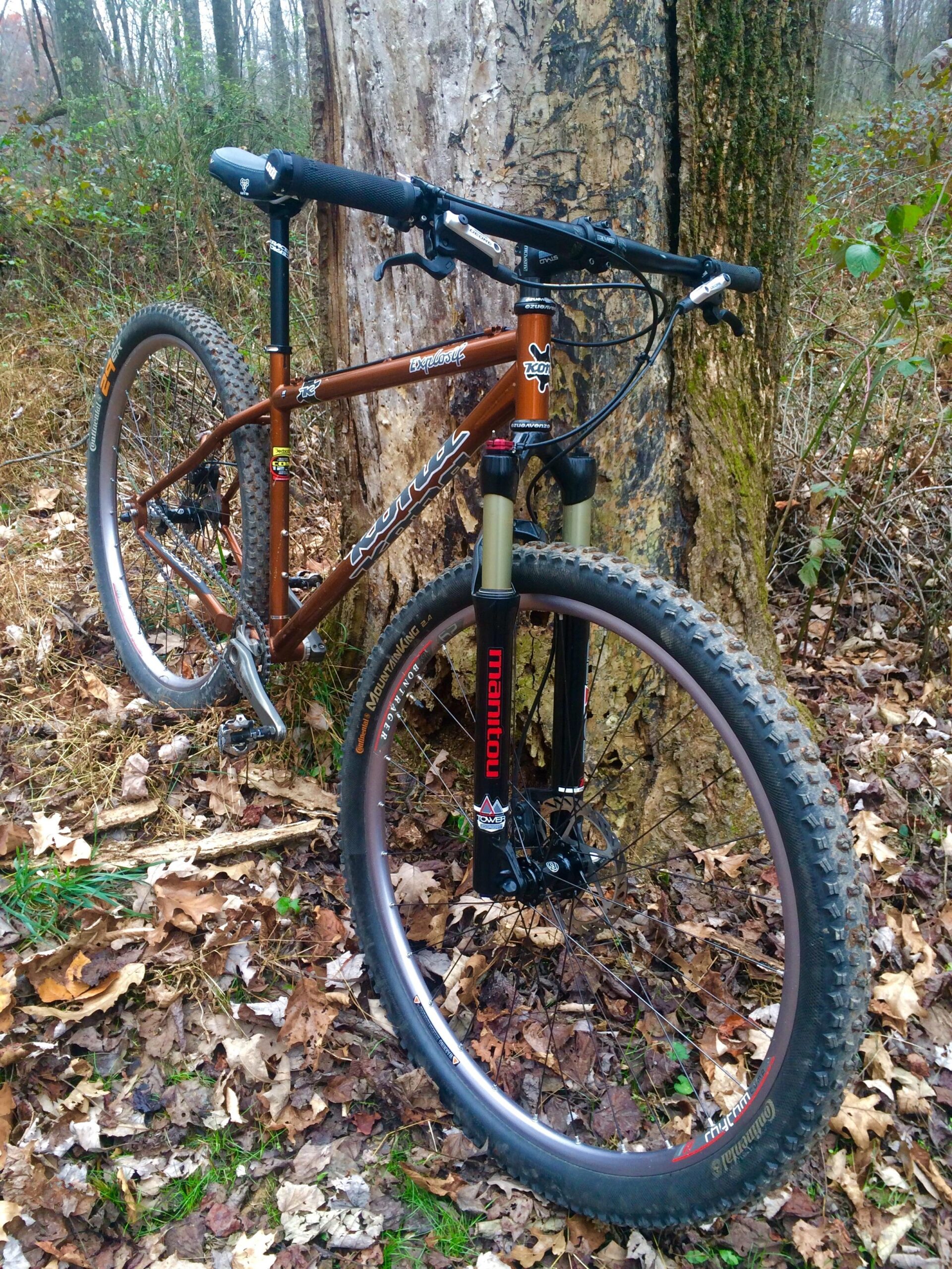 Kona Explosif: A mountain bike leaning against a tree in a forested area, surrounded by fallen leaves and greenery. The bike features a brown frame, suspension fork, and thick tires, indicating its suitability for off-road terrain.