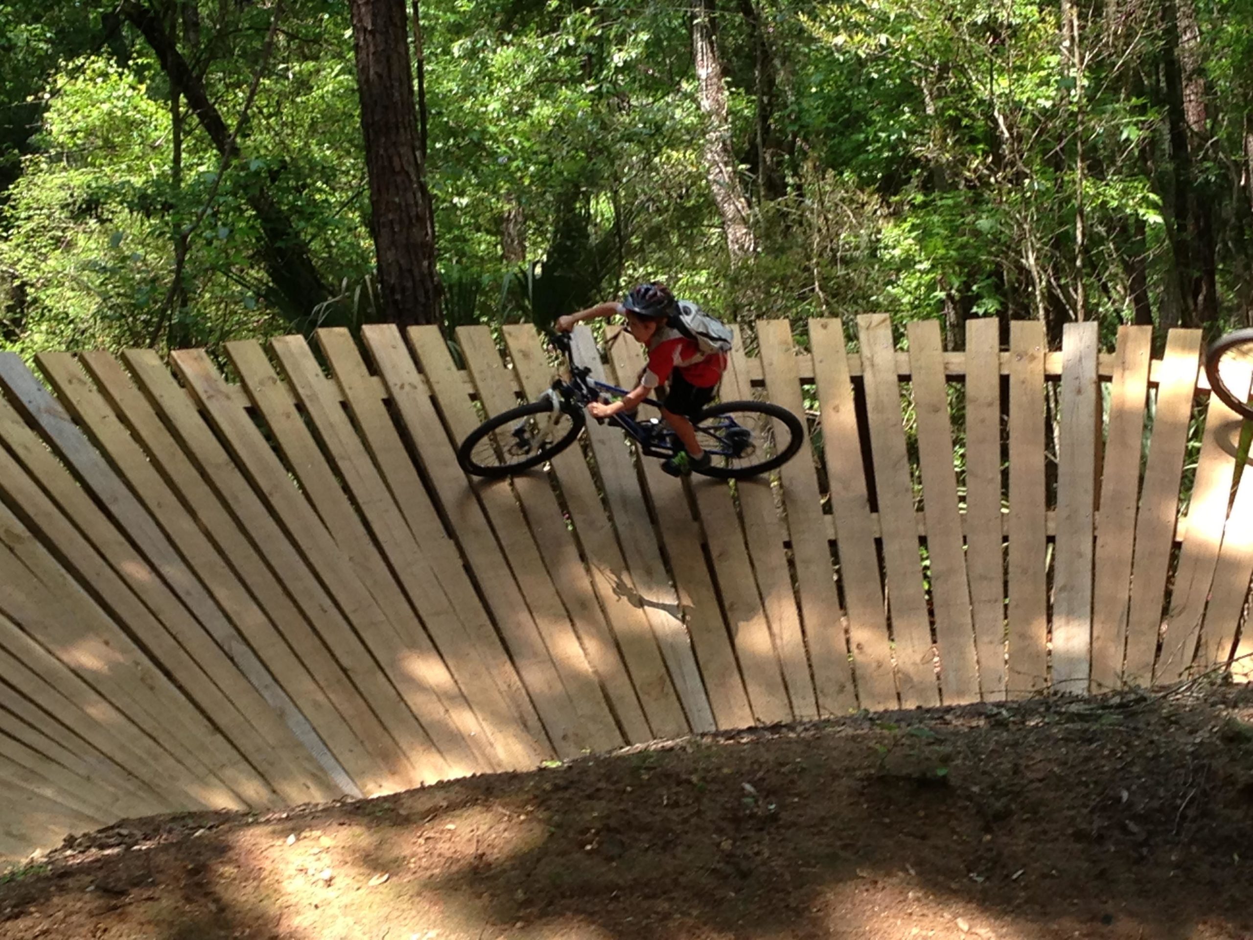 A young cyclist navigating a wooden ramp on a mountain bike trail surrounded by lush greenery. The ramp is built with vertical wooden slats, allowing the rider to maintain speed while performing a lean turn. The scene captures the excitement of outdoor biking in a natural setting. Santos mountain bike trail.