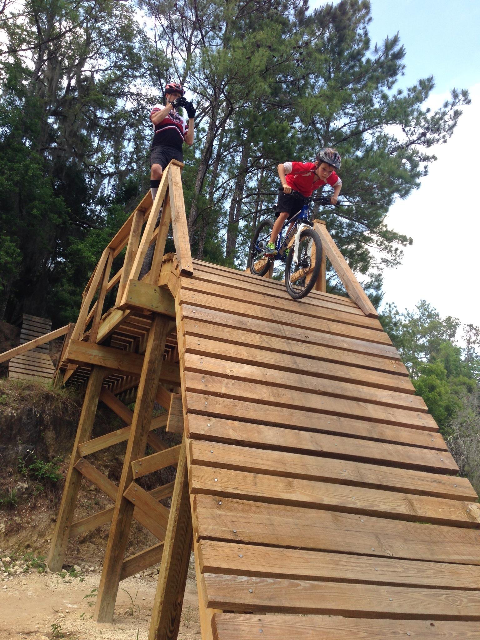A young male cyclist wearing a red shirt and helmet is descending a wooden ramp designed for mountain biking, while another individual in a black and white jersey and helmet stands at the top, holding a camera. The background features trees and a clear sky, emphasizing an outdoor setting. Santos mountain bike trail.