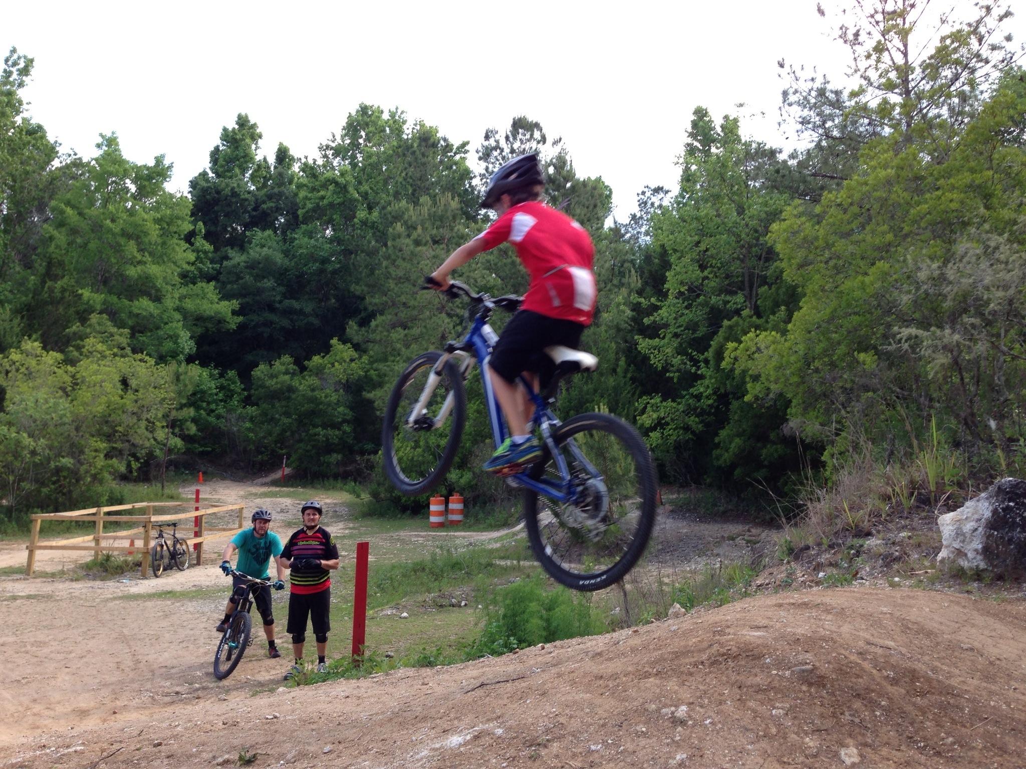 A young cyclist in a red shirt performs a jump on a mountain bike over a dirt mound, while two other cyclists watch in the background. The scene is set in a green, wooded area with dirt trails and obstacles for biking. Santos mountain bike trail.