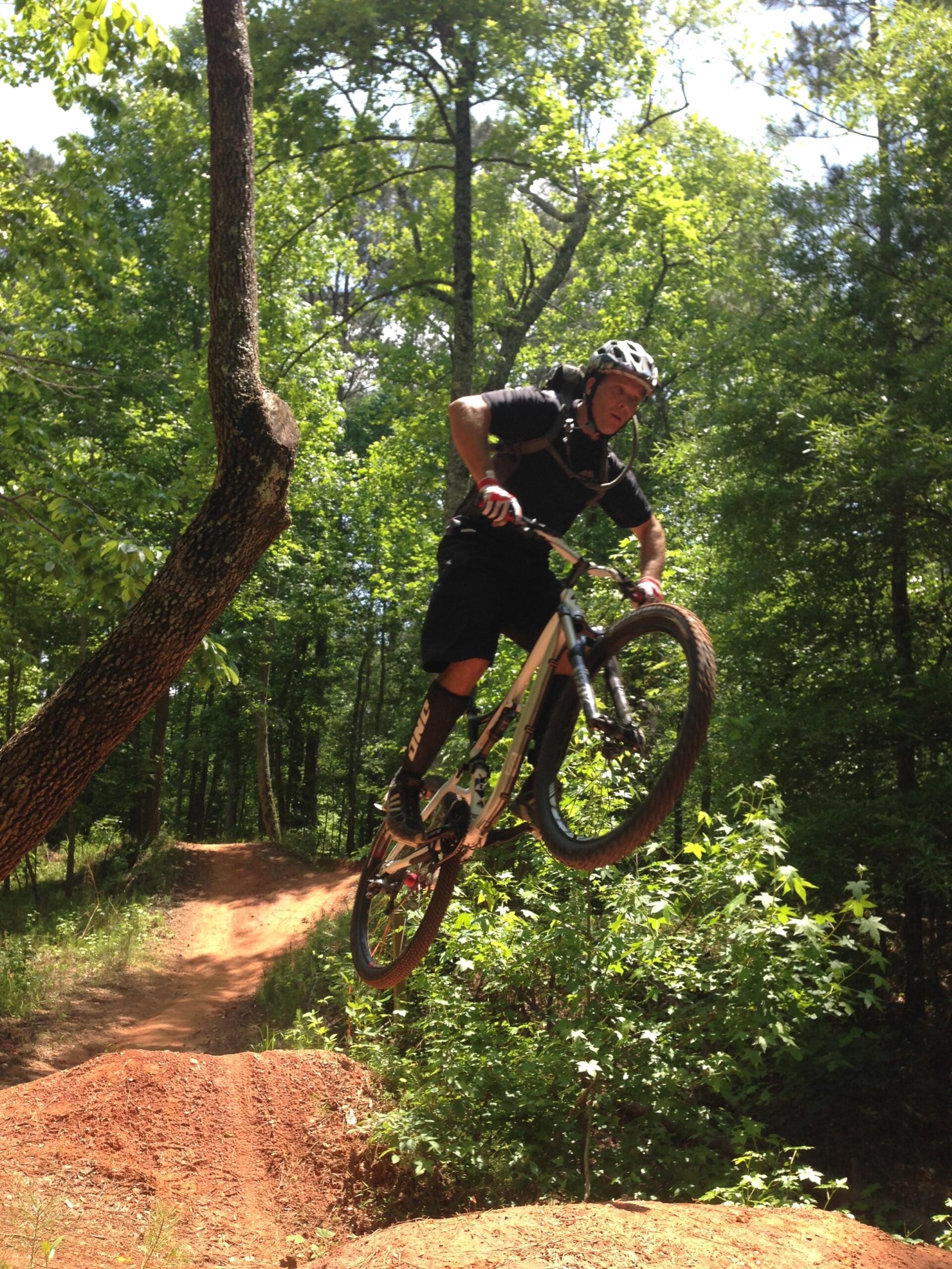 A mountain biker wearing a helmet and protective gear is performing a jump on a dirt track surrounded by trees. The biker is mid-air above a dirt ramp, showcasing an action-packed moment in a forested trail. Blankets Creek mountain bike trail.