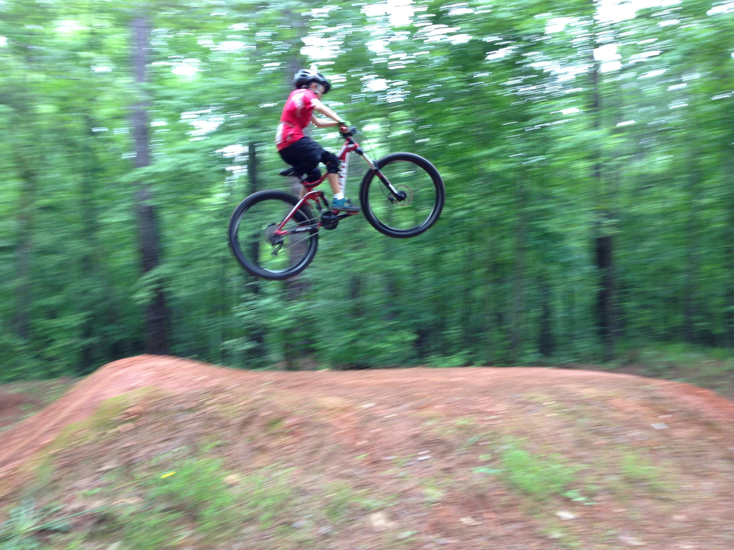 A young cyclist in a red shirt and helmet performs a jump on a mountain bike over a dirt ramp in a wooded area, captured in motion with a blurred background. Blankets Creek mountain bike trail.