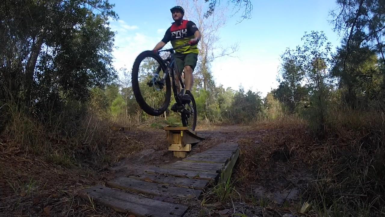 A mountain biker performing a jump on a wooden ramp in a forested area. The rider is wearing a helmet and colorful cycling gear, with one wheel lifted off the ground while approaching the ramp. Surrounding greenery and a clear sky are visible in the background. Nocatee mountain bike trail.