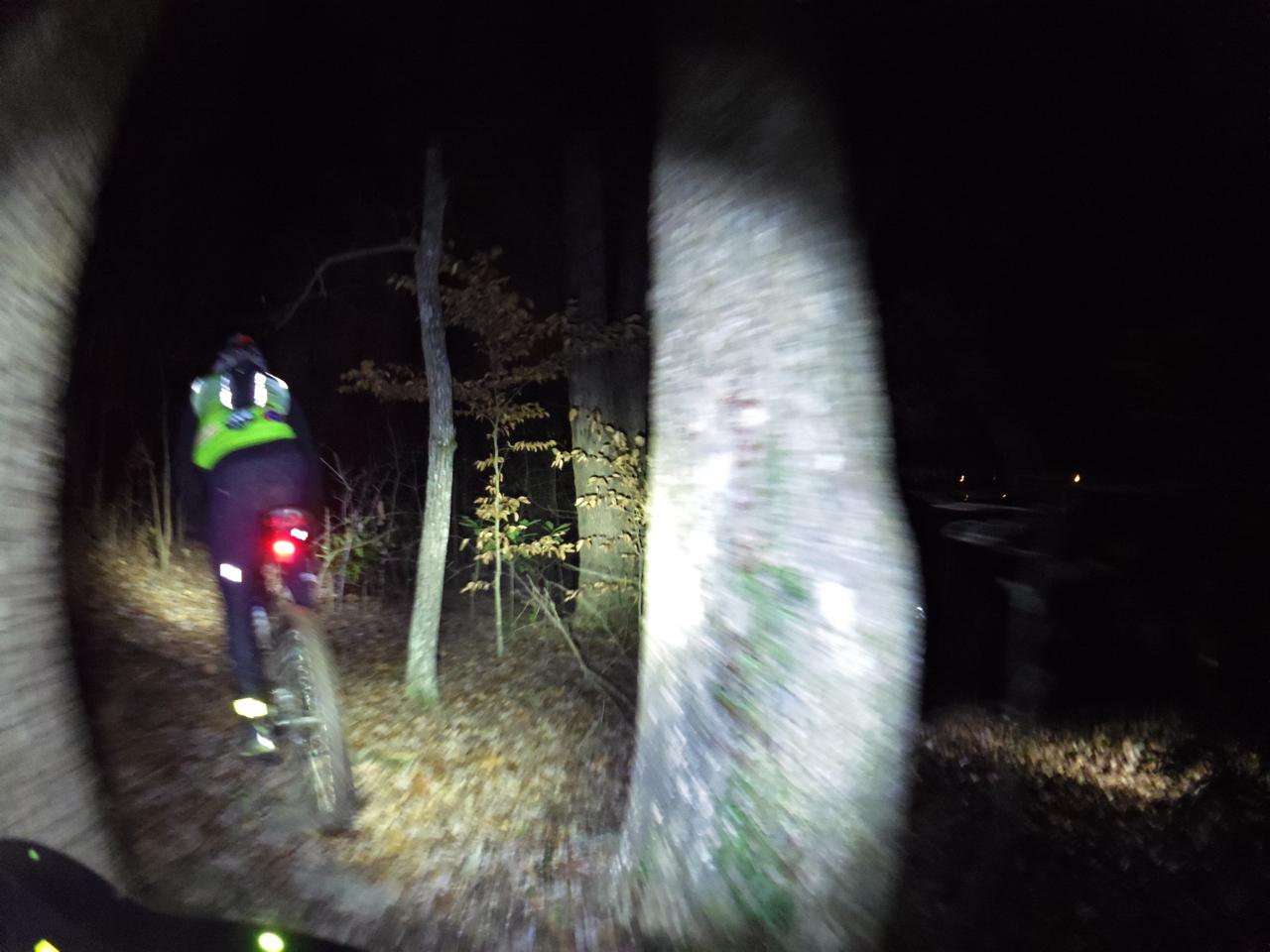 A cyclist riding through a dimly lit forest trail at night, shortly before a tree. The cyclist is wearing a reflective green vest and their bike is equipped with a rear red light. The scene features trees and fallen leaves, illuminated by the bike's headlight. Waterworks / Mason Mill mountain bike trail.