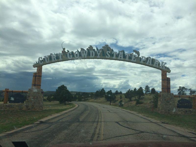 "Archway sign for Curt Gowdy State Park at the entrance, with a winding road leading through a scenic landscape of trees and cloudy skies." Curt Gowdy State Park mountain bike trail.