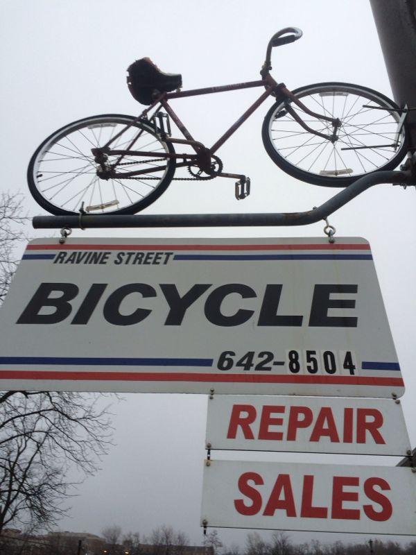 A vintage bicycle hanging above a sign that reads "Ravine Street BICYCLE" with the phone number "642-8504." Below, additional signs state "REPAIR" and "SALES," set against a cloudy sky.