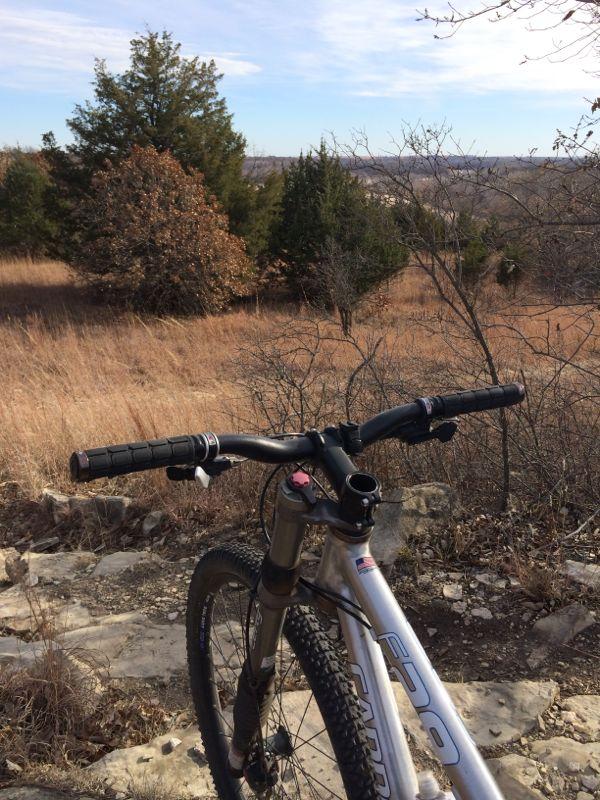 A mountain bike is parked on rocky terrain, with the handlebars and front wheel in focus. In the background, a scenic view of rolling hills and sparse trees under a clear sky can be seen, with tall grass swaying gently in the breeze. Camp Horizon mountain bike trail.