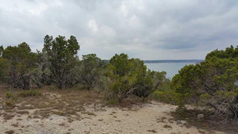 A scenic view of a landscape featuring scattered shrubs and trees, with a cloudy sky above and a body of water visible in the background. The terrain is rocky and dry, typical of a natural outdoor setting. Madrone Trail mountain bike trail.