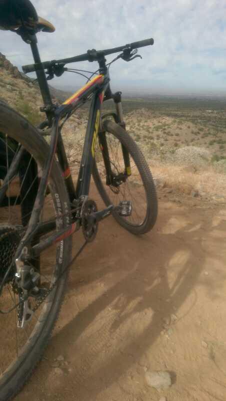 A close-up view of a mountain bike resting on a dirt trail, with a scenic landscape in the background featuring rolling hills and scattered vegetation under a partly cloudy sky. San Tan mountain bike trail.