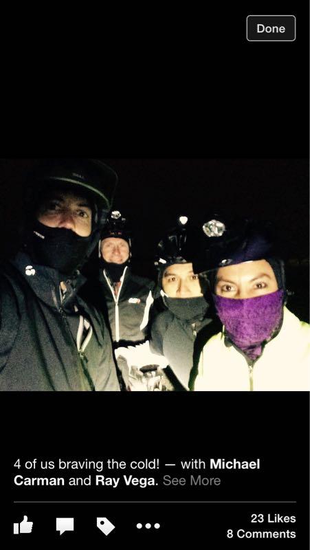 A group of four individuals wearing winter cycling gear and helmets, standing together in a dimly lit outdoor setting. They are bundled up against the cold, with several wearing face coverings and bright reflective materials. The atmosphere conveys camaraderie and determination to cycle despite the chilly conditions. Slaughter Pen Trail mountain bike trail.