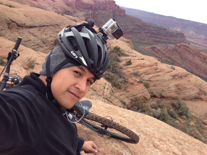 A person wearing a helmet and a black jacket takes a selfie while sitting on rocky terrain, with a bicycle nearby. In the background, a scenic view of cliffs and a winding river is visible under a cloudy sky. A camera is mounted on the helmet, suggesting an adventure or outdoor activity. Slickrock mountain bike trail.