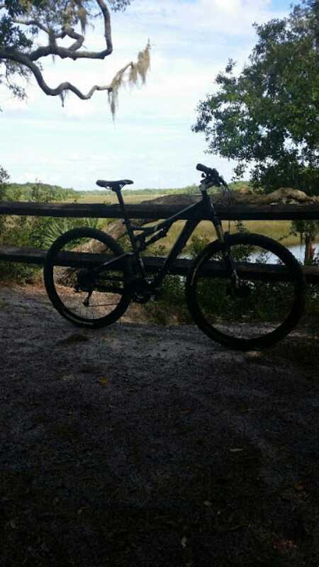 A black mountain bike parked near a wooden fence, with a scenic view of a river and trees in the background under a partly cloudy sky. Spruce Creek Preserve mountain bike trail.