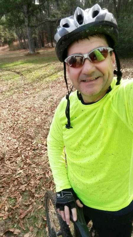 A person wearing a bright yellow long-sleeve shirt and a black helmet is smiling in a wooded area, holding a mountain bike. The ground is covered with leaves, and there are visible bike tracks in the background. The individual is also wearing sunglasses and gloves, indicating they are engaged in outdoor cycling. Long Cane Horse Trail mountain bike trail.