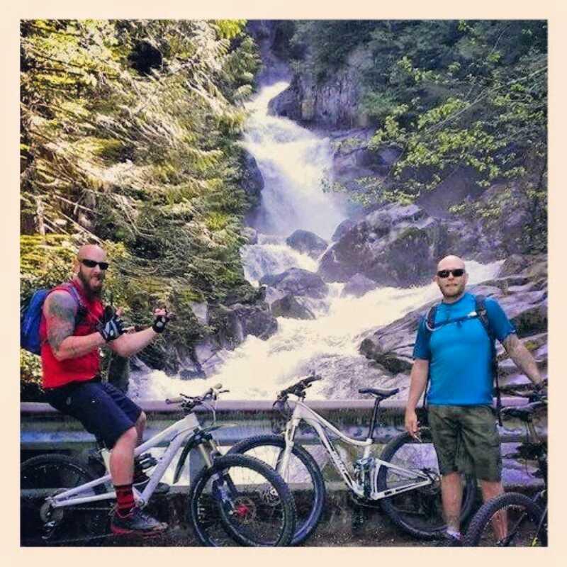 Two mountain bikers pose beside their white bicycles in front of a scenic waterfall surrounded by lush greenery. One biker, wearing a red tank top and shorts, raises his arm playfully, while the other, dressed in a blue shirt and shorts, stands confidently with a slight smile. The cascading water and rocky landscape add to the adventure-filled atmosphere of the scene. Middle Fork Snoqualmie mountain bike trail.