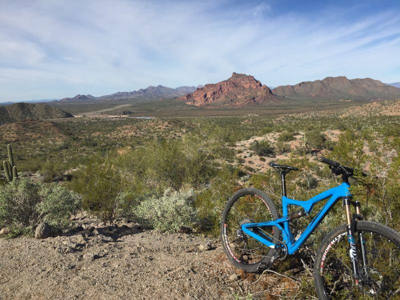 A blue mountain bike rests on rocky terrain, overlooking a vast desert landscape filled with green vegetation and cacti. In the distance, red rocky formations rise against a clear blue sky, suggesting a scenic outdoor location perfect for biking and exploration. Hawes Loop mountain bike trail.