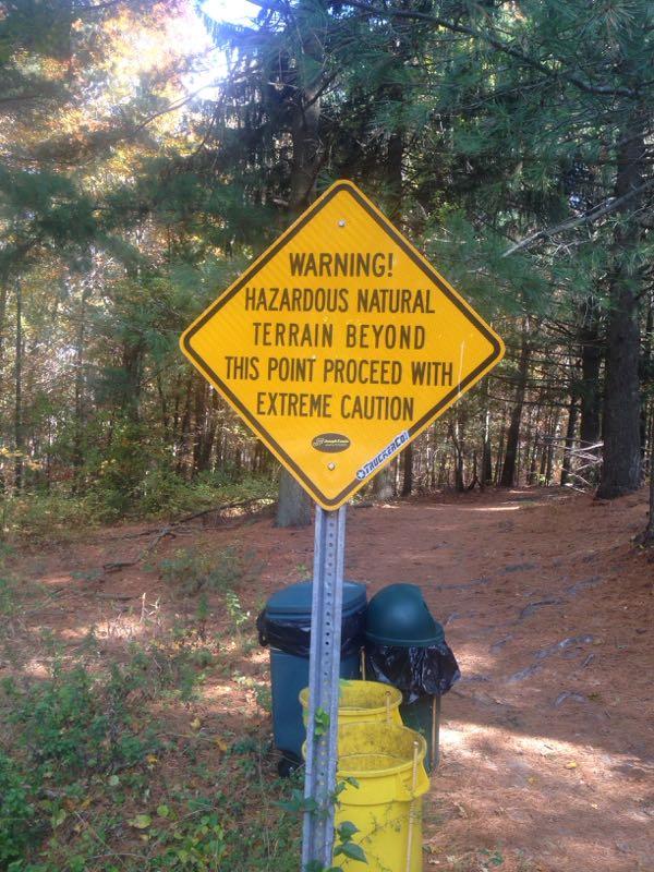 A yellow warning sign indicating hazardous natural terrain ahead, with instructions to proceed with extreme caution. The sign is mounted on a post near trash bins, surrounded by a forested area with pine trees and a carpet of fallen leaves. Cere's Park mountain bike trail.