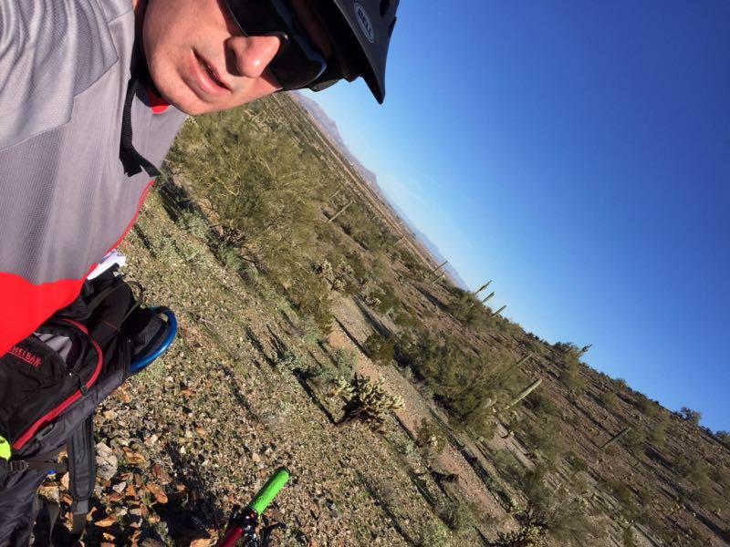 A person wearing a helmet and sunglasses takes a selfie while mountain biking in a desert landscape, surrounded by cacti and rocky terrain under a clear blue sky. A bicycle is partially visible in the foreground. Casa Grande Mountain mountain bike trail.