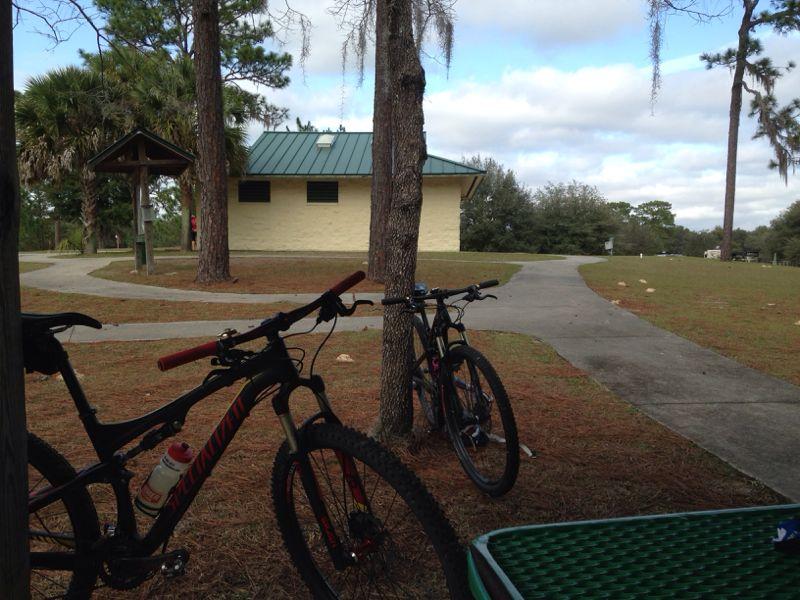 Two mountain bikes are parked on the ground in a wooded area, with a view of a park building with a green metal roof in the background. There are pine trees and a walking path visible, along with a picnic table in the foreground. The sky is partly cloudy, suggesting a pleasant day in a natural setting. Santos mountain bike trail.