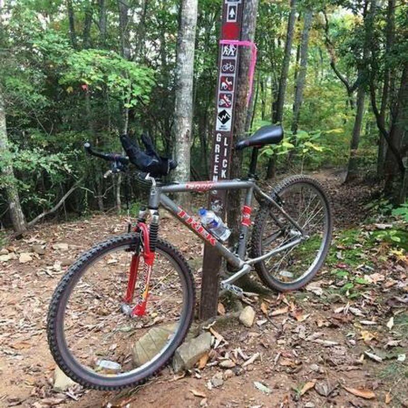 A mountain bike leaning against a trail marker in a wooded area, with various trail signs including difficulty levels and directions. The bike features a gray frame and red front fork, and there's a water bottle attached to the frame. The background shows a tree-filled path with fallen leaves on the ground. Greenslick mountain bike trail.