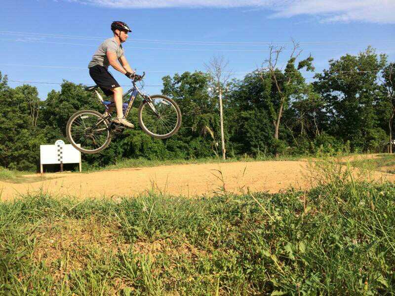A person wearing a helmet and a gray shirt is performing a jump on a mountain bike over a dirt path, with greenery in the background under a clear blue sky. Lester Singhas BMX Track mountain bike trail.