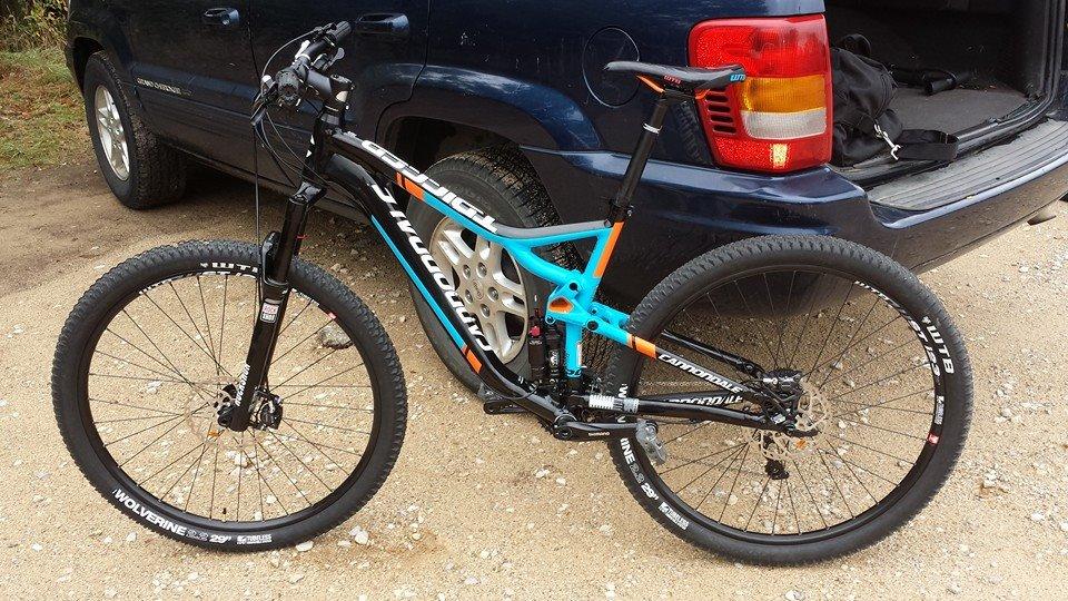 Cannondale Trigger: A mountain bike with a black and blue frame featuring orange accents, positioned on a gravel surface next to a parked dark blue SUV. The bike has oversized tires and front suspension, highlighting its suitability for off-road trails.