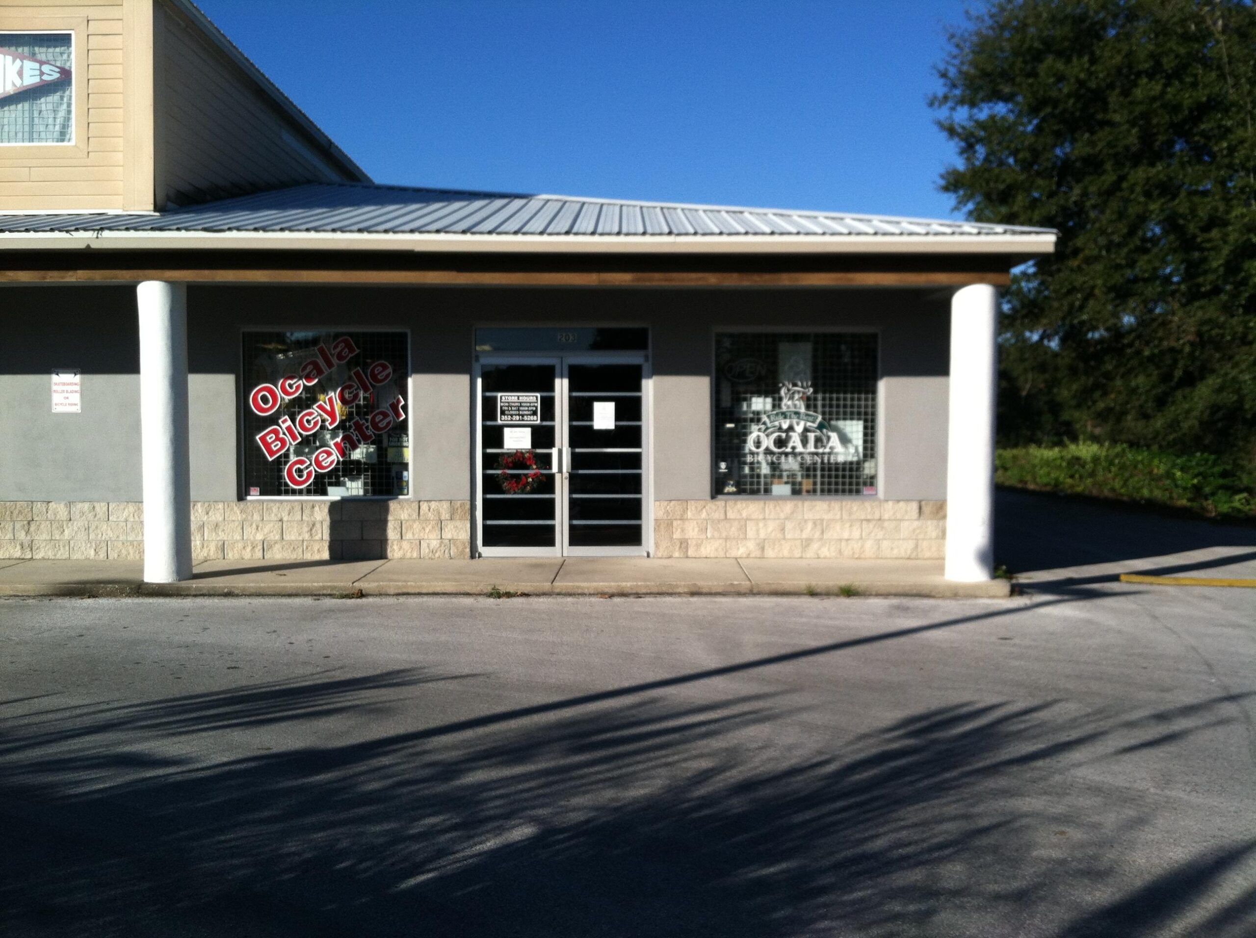 Exterior view of the Ocala Bicycle Center, featuring large windows with signage indicating the business name. The building has a modern design with a metal roof and a concrete foundation. There's a decorative wreath on one of the windows, and a clear blue sky can be seen above.