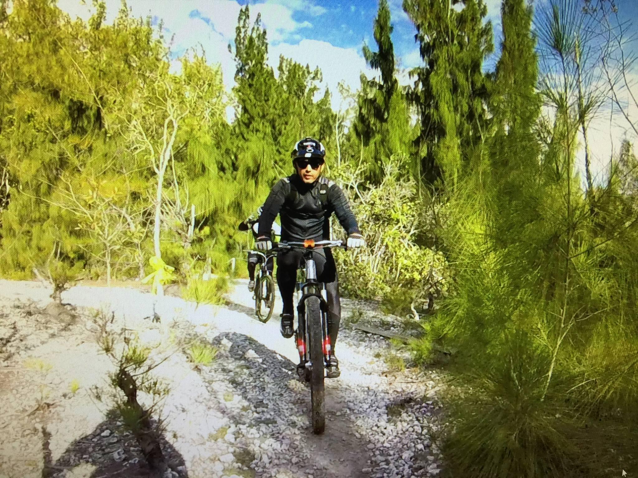A mountain biker navigating a rocky trail through a lush, green forest, with tall trees and shrubs surrounding the path. The cyclist is wearing a black helmet and gear, focused on the trail ahead.