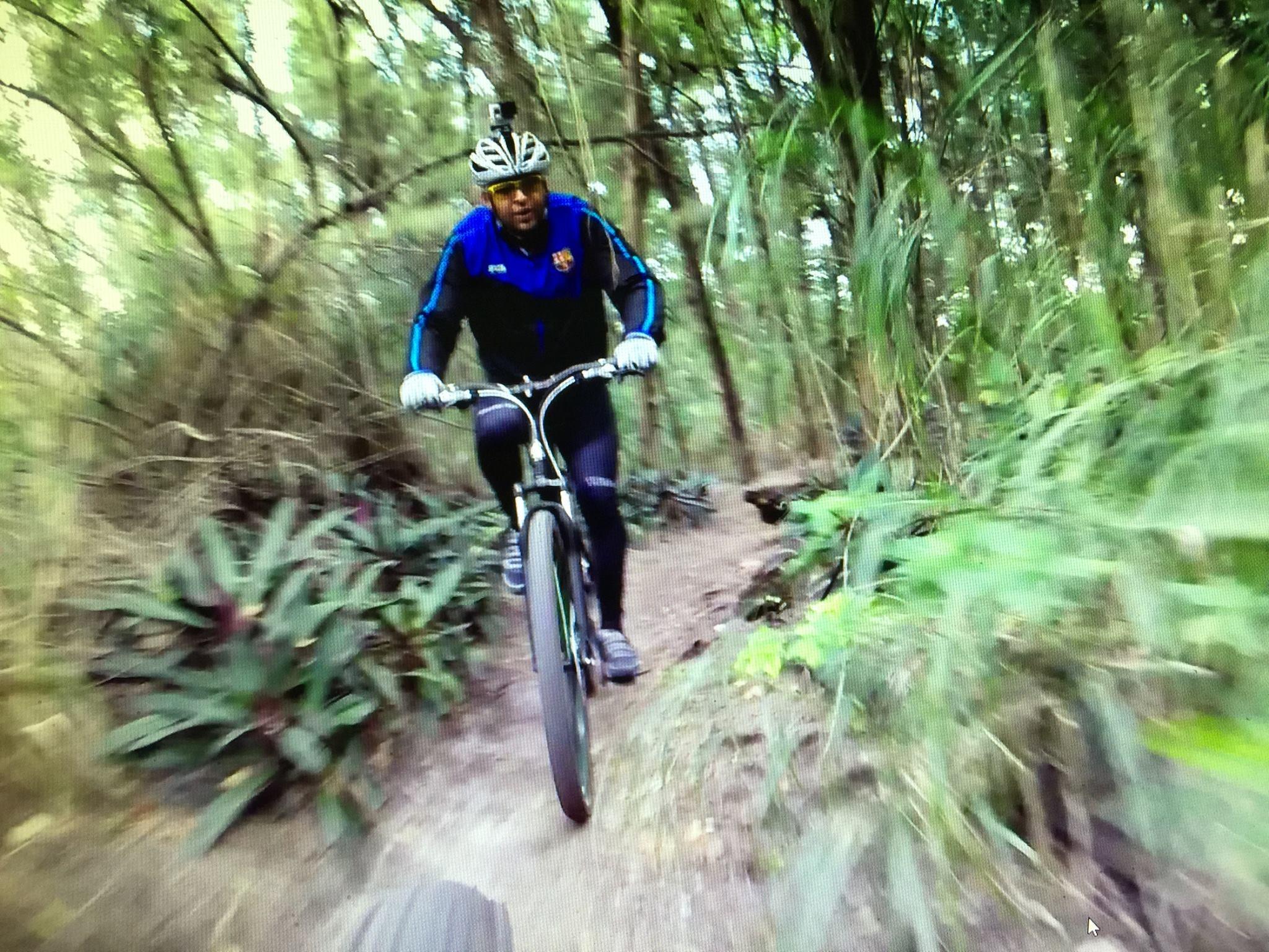 A cyclist navigating a winding dirt trail surrounded by greenery and plants, wearing a helmet with an action camera mounted on top, and dressed in athletic gear.