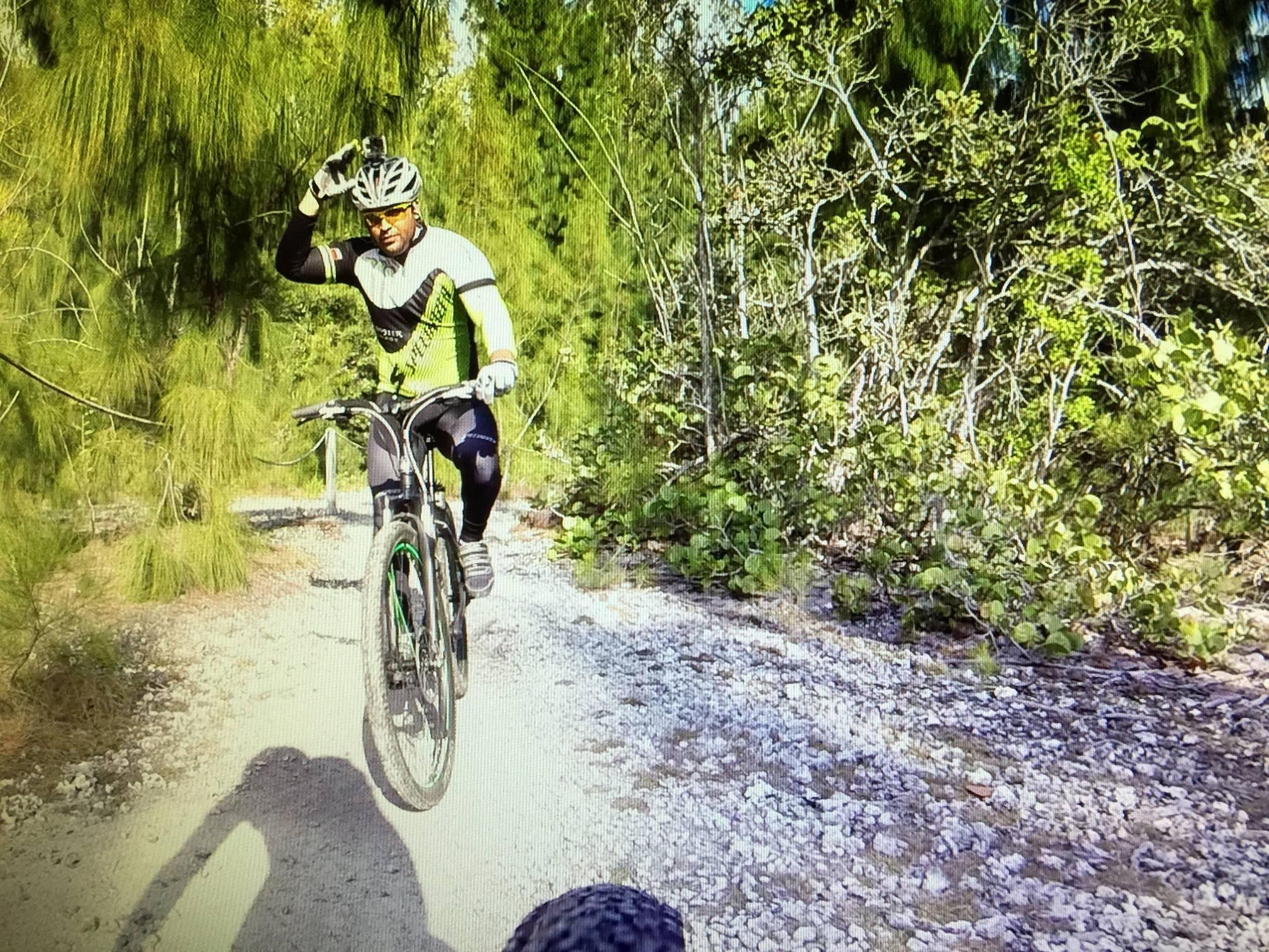 A mountain biker wearing a bright yellow and black jersey and helmet rides down a gravel path surrounded by green foliage. The cyclist raises one hand in a gesture, possibly to greet or signal. Sunlight filters through the trees, highlighting the vibrant surroundings.