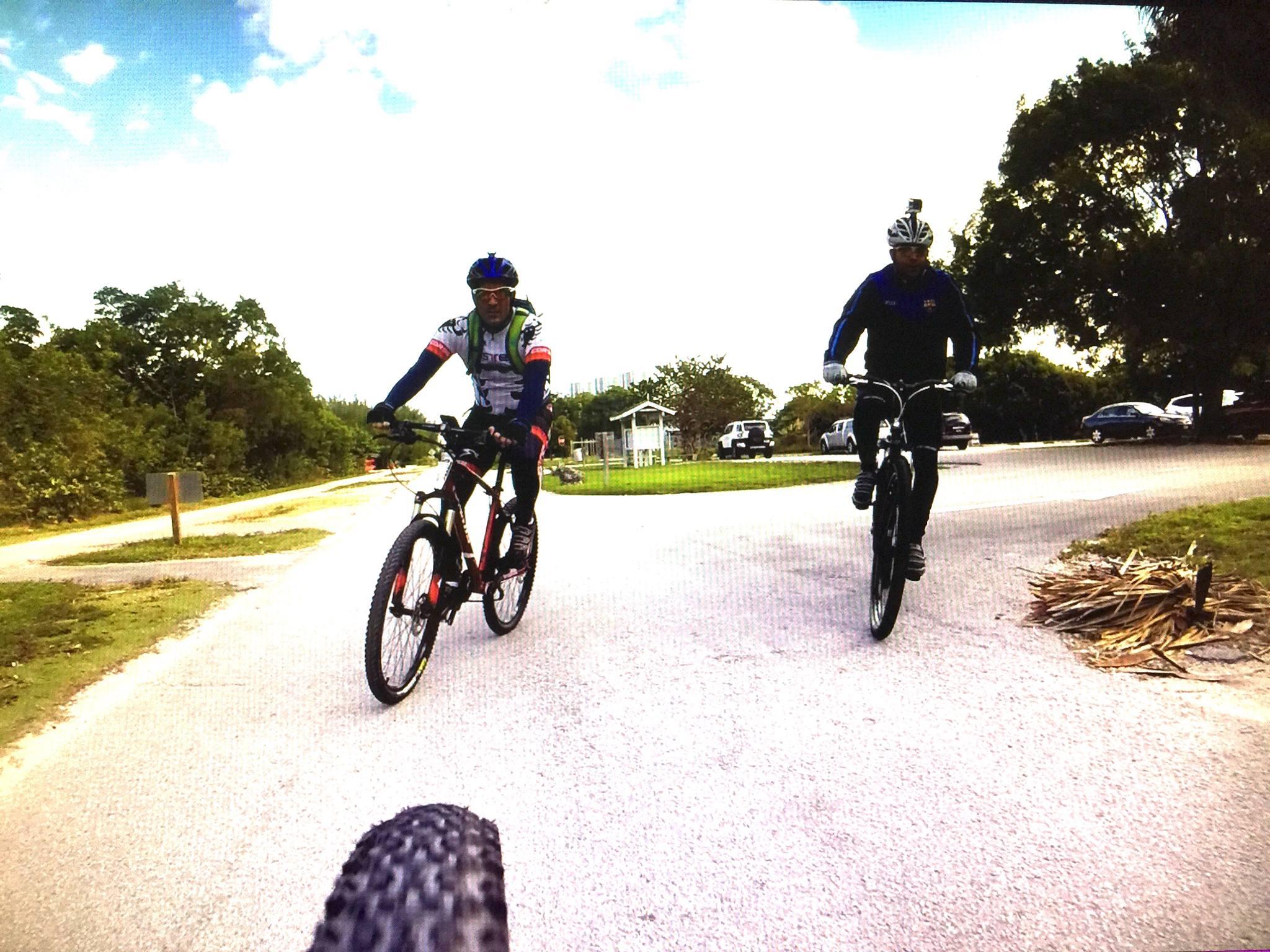 Two cyclists riding on a path in a park. One cyclist is on a red mountain bike, wearing a green helmet and a cycling jersey, while the other cyclist is on a black bike, dressed in a blue jacket and helmet. The background features trees and parked cars, with a clear sky above. Oleta River State Park mountain bike trail.
