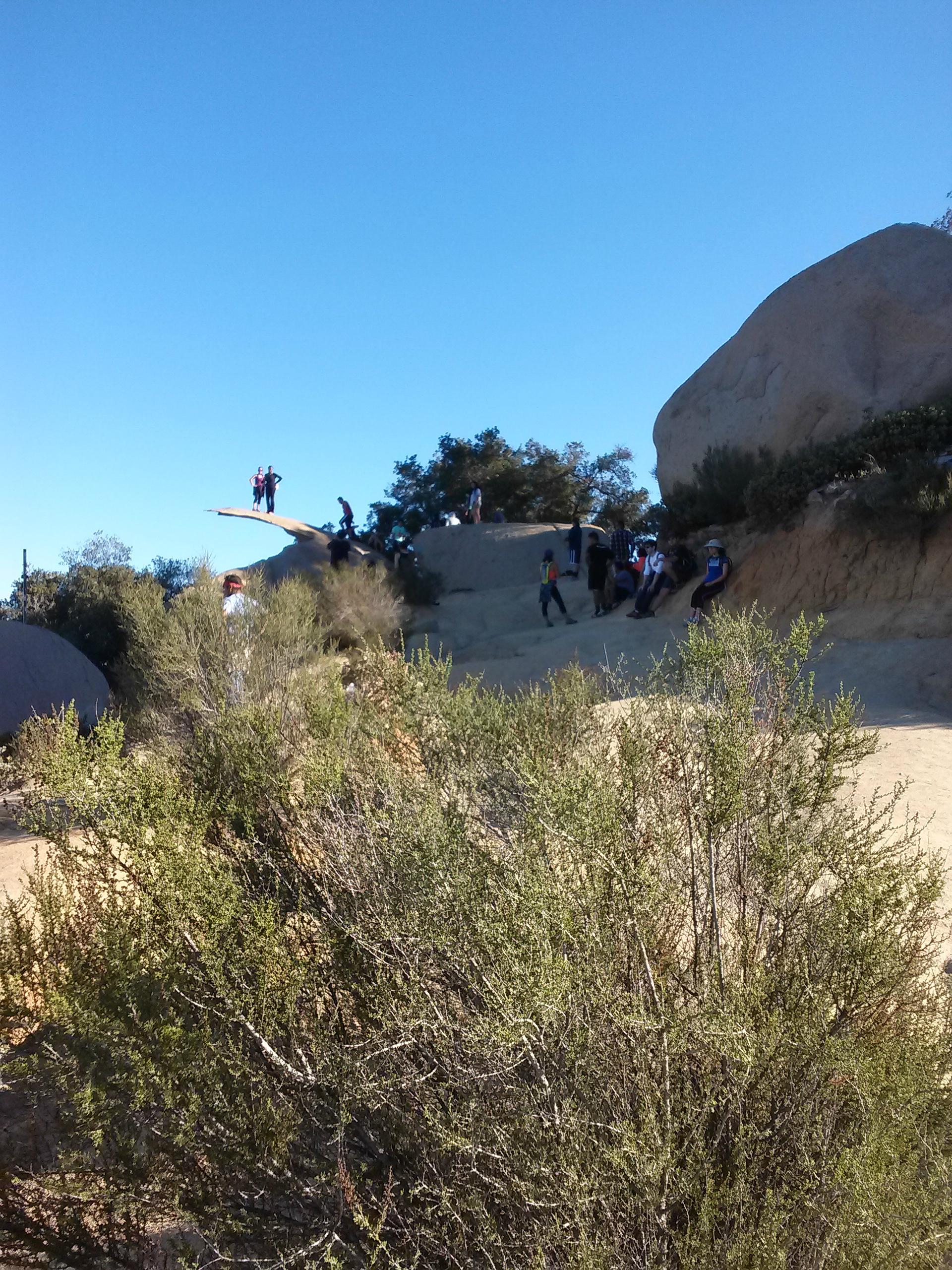 A scenic outdoor landscape featuring a rocky terrain under a clear blue sky. Several people are engaged in recreational activities, including two individuals standing on a prominent rock formation. The foreground includes patches of shrubbery and scattered boulders. Fry-Koegel Trail mountain bike trail.