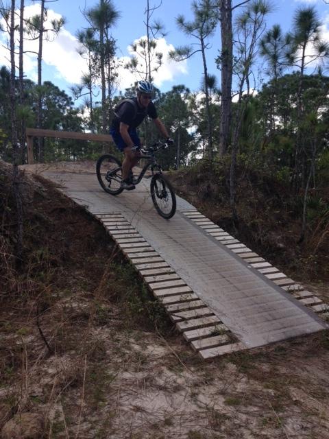 A mountain biker riding down a wooden ramp through a forested area, surrounded by tall pine trees and blue skies. The cyclist wears a helmet and a backpack, showcasing a dynamic action shot in an outdoor setting. Halpatiokee mountain bike trail.