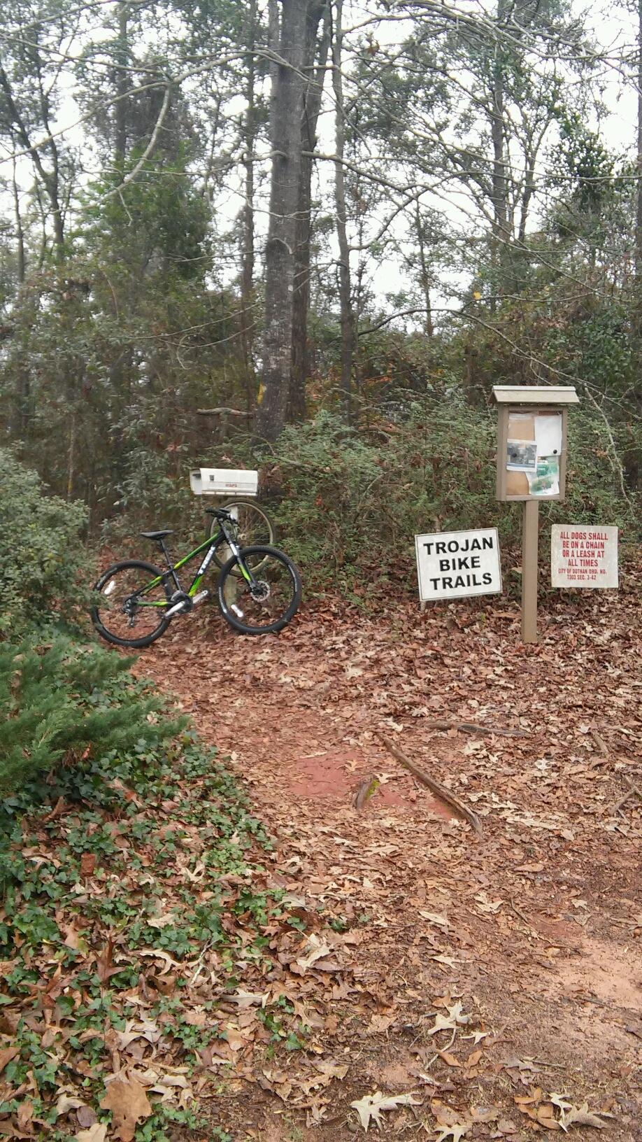 Trek Marlin 6: A mountain bike resting against a white mailbox near a trail sign that reads "Trojan Bike Trails." The area is surrounded by trees and bushes, and the ground is covered with fallen leaves. There is also an informational board and a sign noting dog leash regulations.