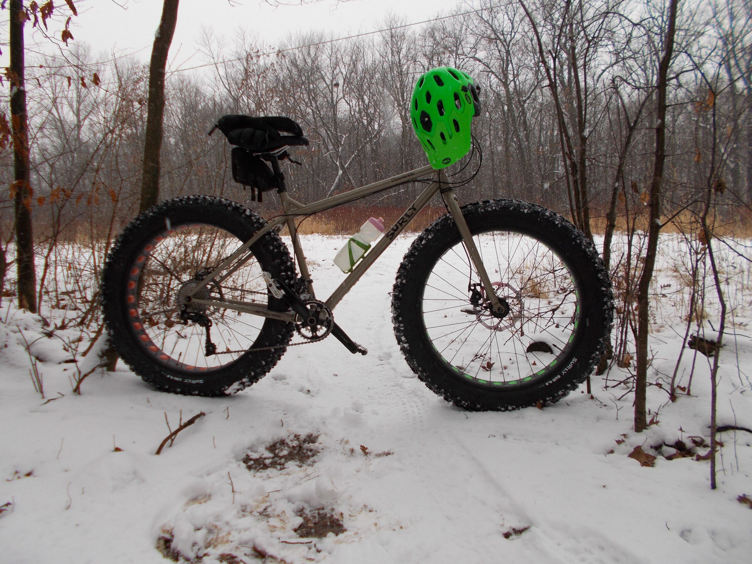 Surly Moonlander: A fat bike with oversized tires is parked on a snowy trail, surrounded by bare trees. A bright green helmet rests on the bike's frame, and the ground is covered in fresh snow. The scene is overcast and wintery, indicating cold weather conditions.