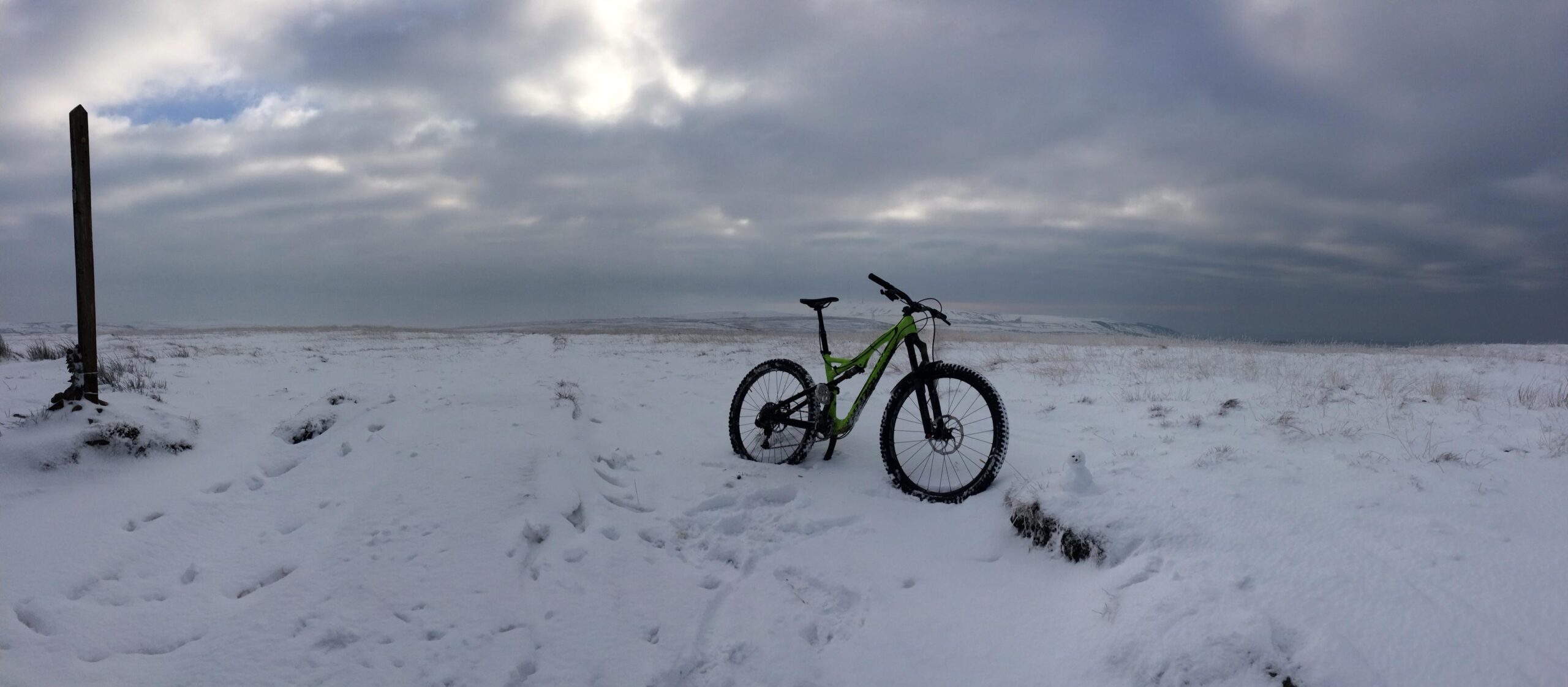 Specialized Stumpjumper FSR Expert Carbon Evo 650B: A mountain bike with a green frame rests on a snowy landscape, with a wooden signpost in the background and a small snowman nearby. The scene is set under overcast skies, showcasing a winter wilderness.
