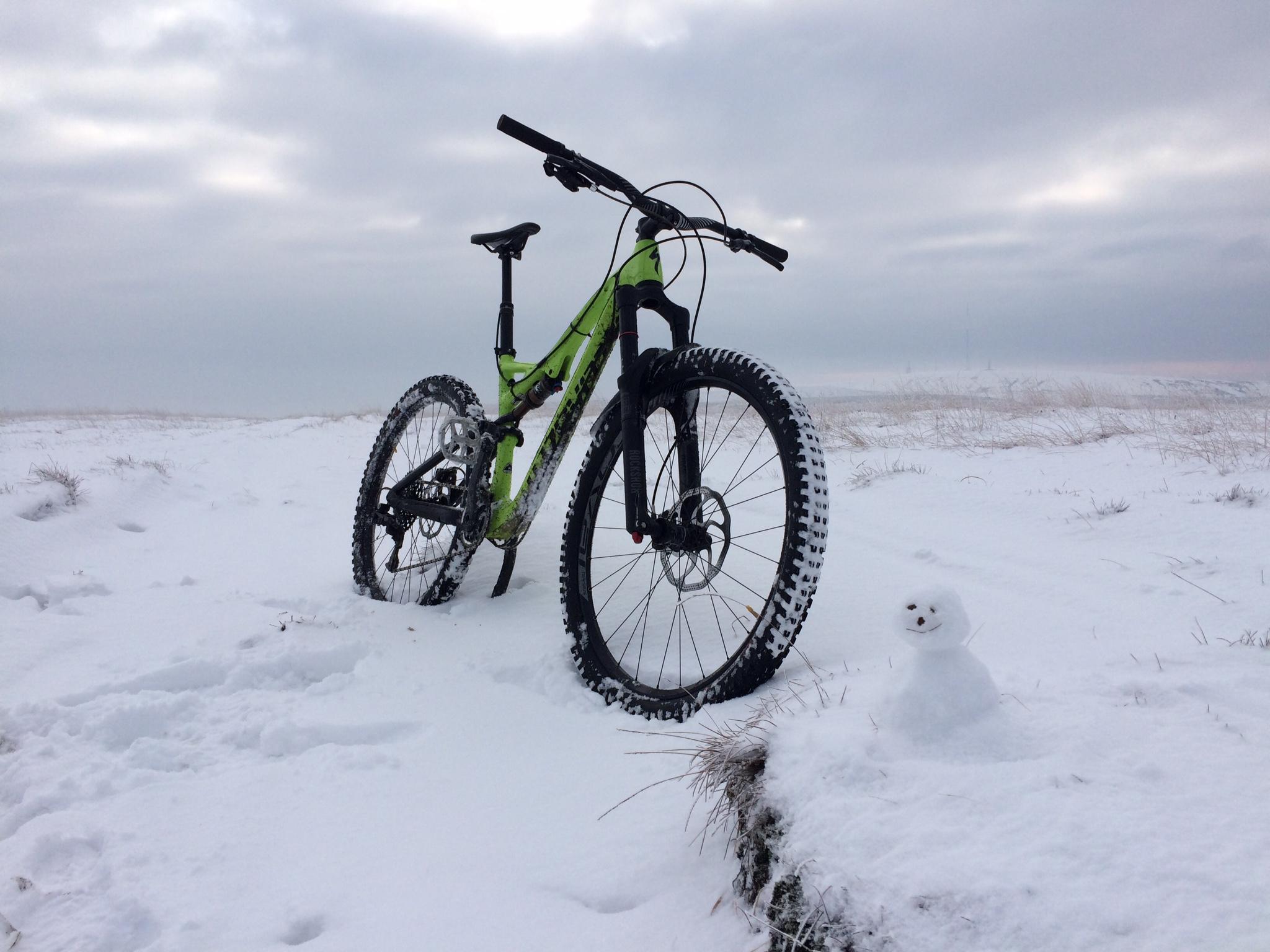 Specialized Stumpjumper FSR Expert Carbon Evo 650B: A mountain bike with a bright green frame is parked in a snowy landscape, with a small snowman nearby. The scene features overcast skies, and patches of grass are visible beneath the snow. The bike is positioned prominently, showcasing its large tires, while the snowman adds a playful touch to the wintery setting.
