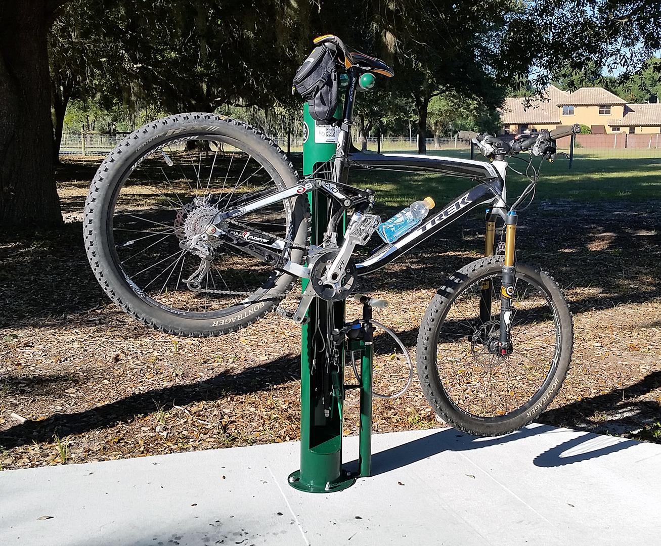 A black and silver mountain bike is mounted on a green bike repair stand in a park. A water bottle is secured to the bike, and the background features trees and a distant building. The bike