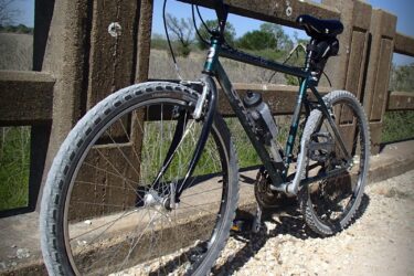 A mountain bike leaning against a textured wooden railing, positioned on a gravel path. The bike features a green frame, wide tires, and a water bottle mounted on the frame, with a natural landscape of tall grass and trees in the background under a clear blue sky.