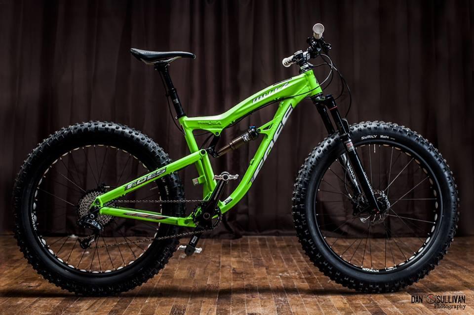 Foes Mutz: A vibrant green mountain bike is displayed against a dark backdrop, showcasing its sleek design and robust tires. The bike features a lightweight frame, wide tires with deep treads, and suspension components, emphasizing its suitability for off-road trails. The wooden floor adds a rustic touch to the overall composition.