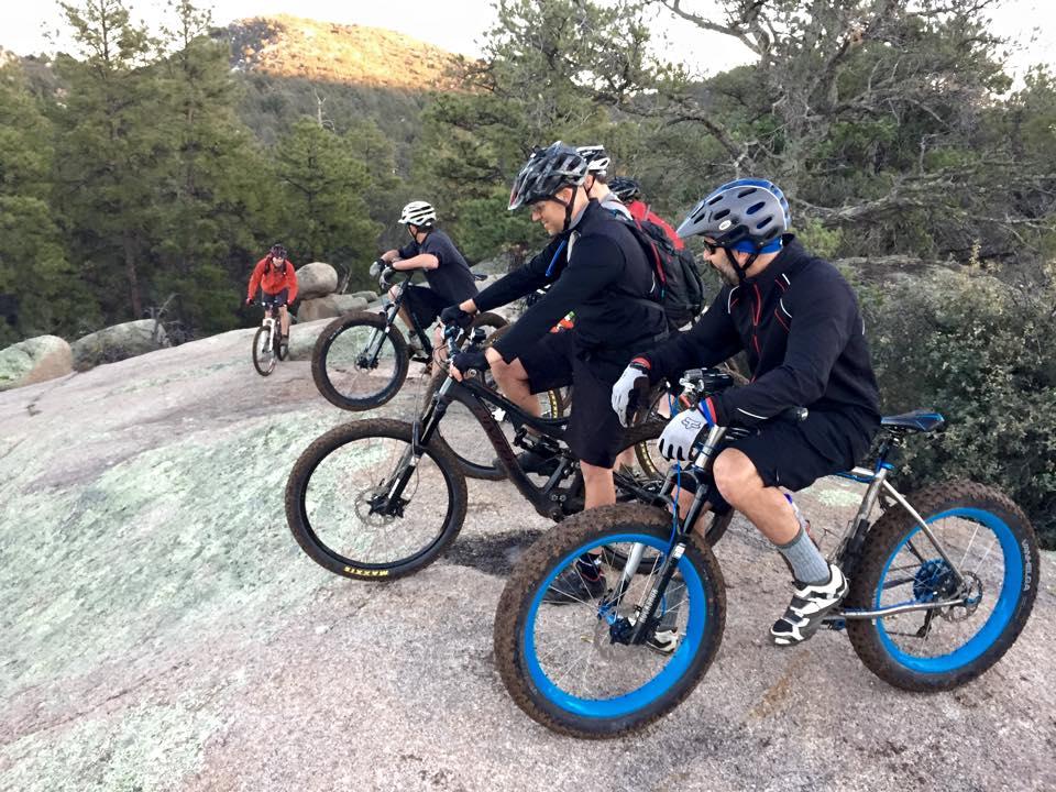 A group of five mountain bikers navigating rocky terrain, dressed in cycling gear and helmets. The scene is set in a natural outdoor environment, surrounded by trees and hills in the background, highlighting an adventurous biking activity. Granite Dells mountain bike trail.