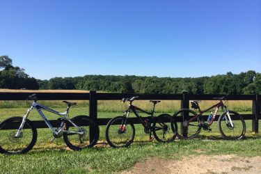 Jamis Exile Comp: Three mountain bikes are leaning against a wooden fence in a scenic outdoor setting. The background features a lush green field and trees under a clear blue sky. The sun is shining, creating a bright and inviting atmosphere.