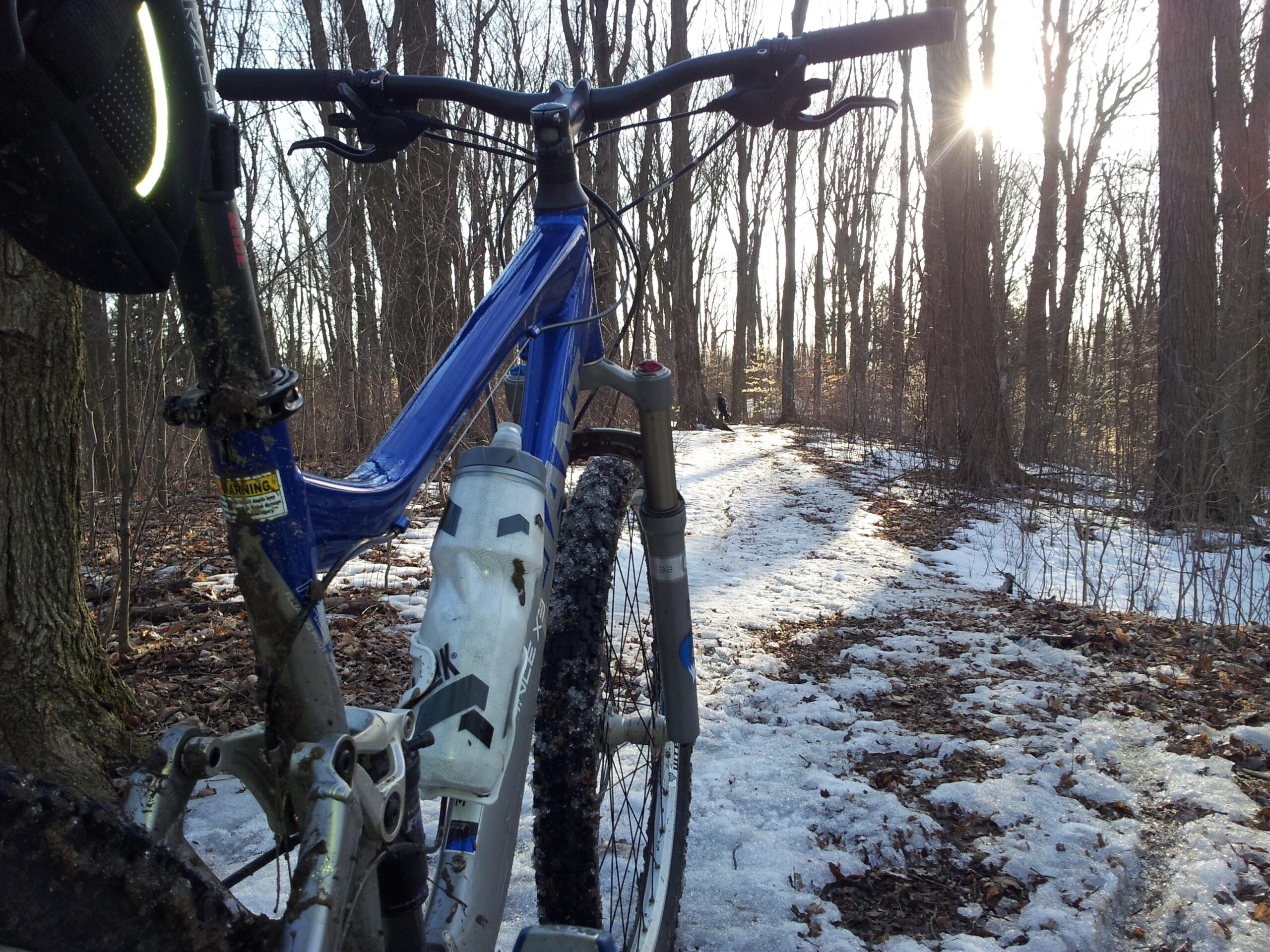 Giant Trance X3: A close-up view of a blue mountain bike with a water bottle mounted on the frame, parked on a snowy dirt trail surrounded by trees. The sun is shining through the branches, and the trail shows a mix of snow and fallen leaves, suggesting a late winter or early spring setting.