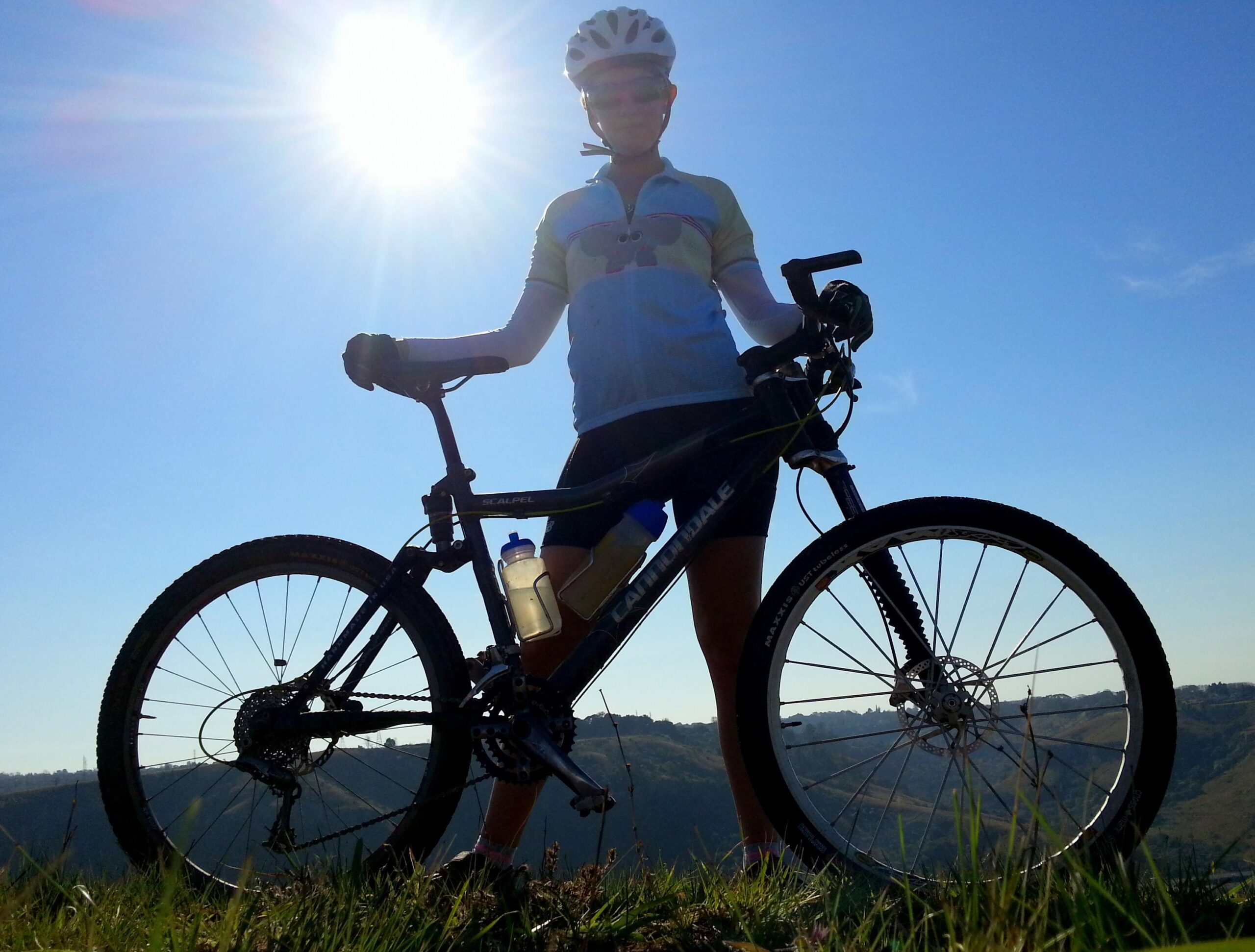 Cannondale Scalpel: A silhouette of a cyclist standing next to a mountain bike on a grassy hill, with the sun shining brightly in the background against a clear blue sky. The cyclist is wearing a light blue cycling jersey and shorts, and a water bottle is attached to the bike.