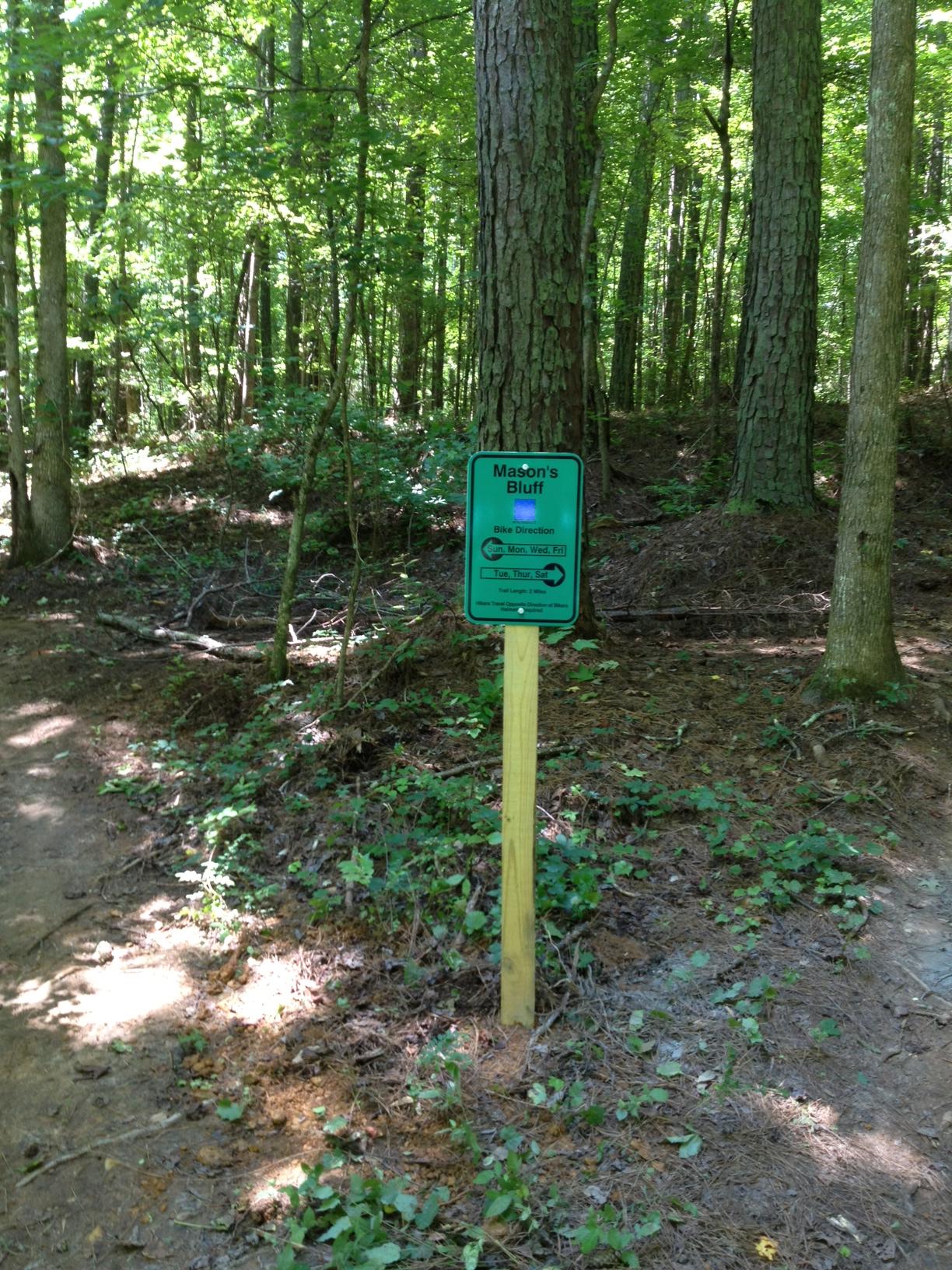 A signpost in a wooded area indicating the bike direction for Mason's Bluff trail. The sign features a green background with directional arrows and guidelines for mountain biking. Tall trees and lush greenery surround the path, which is partially covered with pine needles. Mason's Bluff mountain bike trail.
