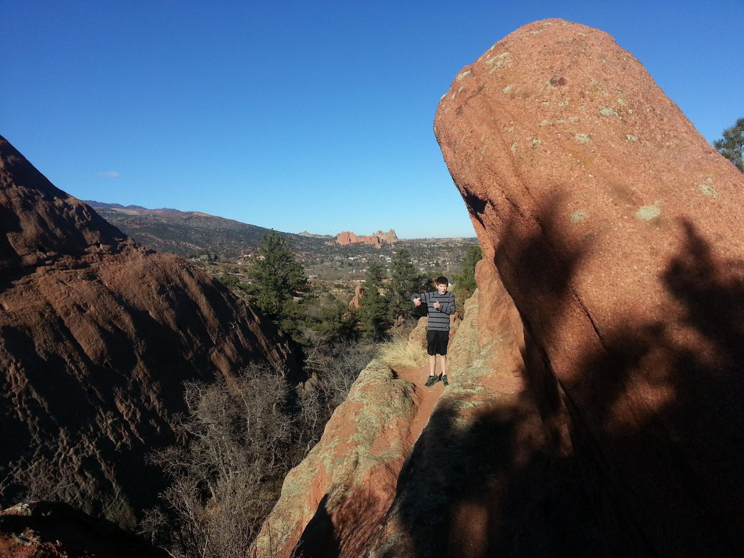 A young person stands on a narrow path between large red rock formations, surrounded by a scenic landscape featuring distant mountains and greenery under a clear blue sky. Red Rock Canyon mountain bike trail.