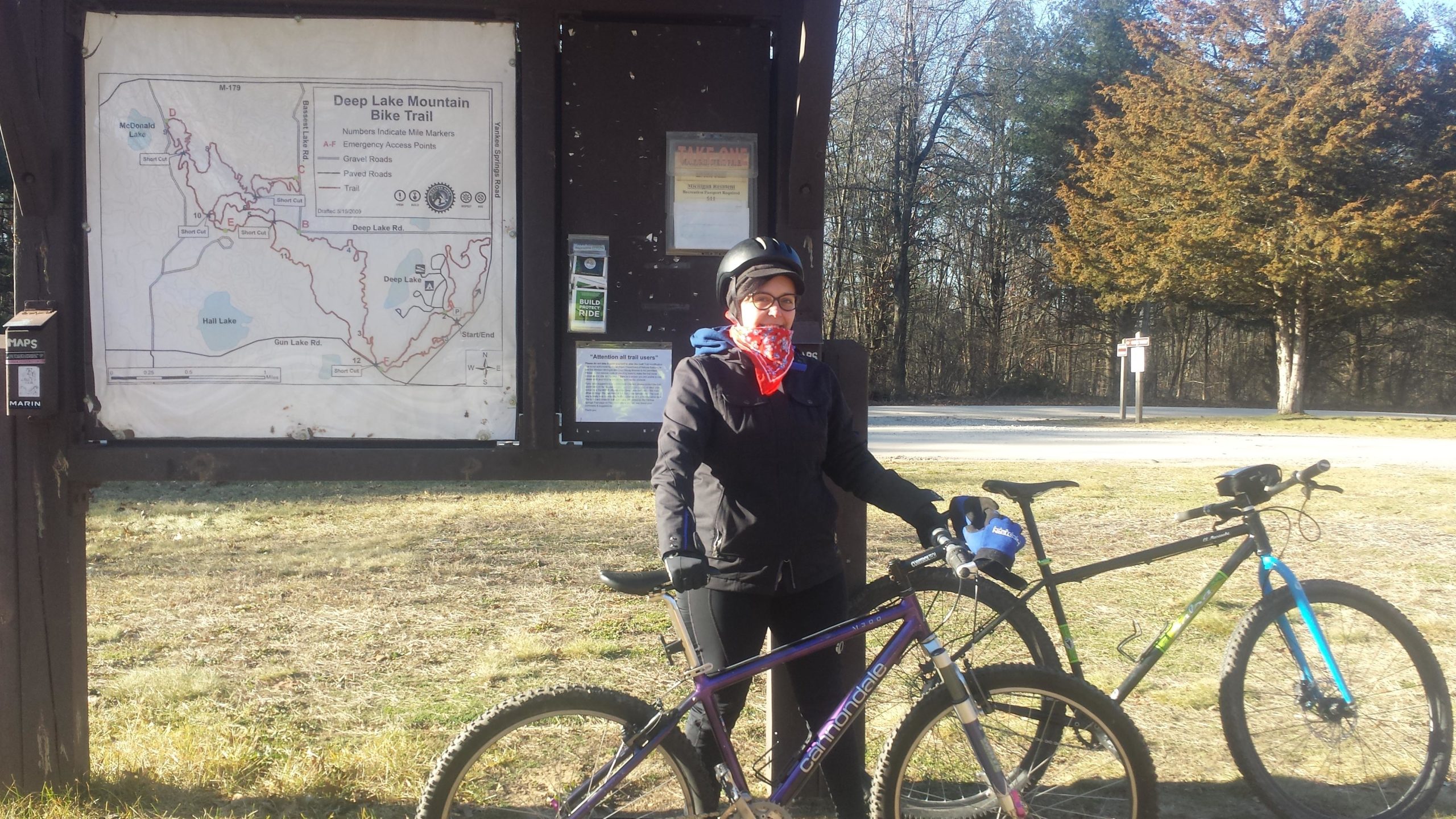 A person wearing a black jacket and a red bandana stands beside a mountain bike in front of a trail map sign for the Deep Lake Mountain Bike Trail. The background features a wooded area with trees, and another bike is parked nearby. The setting appears to be a sunny day in a natural outdoor location. Yankee Springs mountain bike trail.