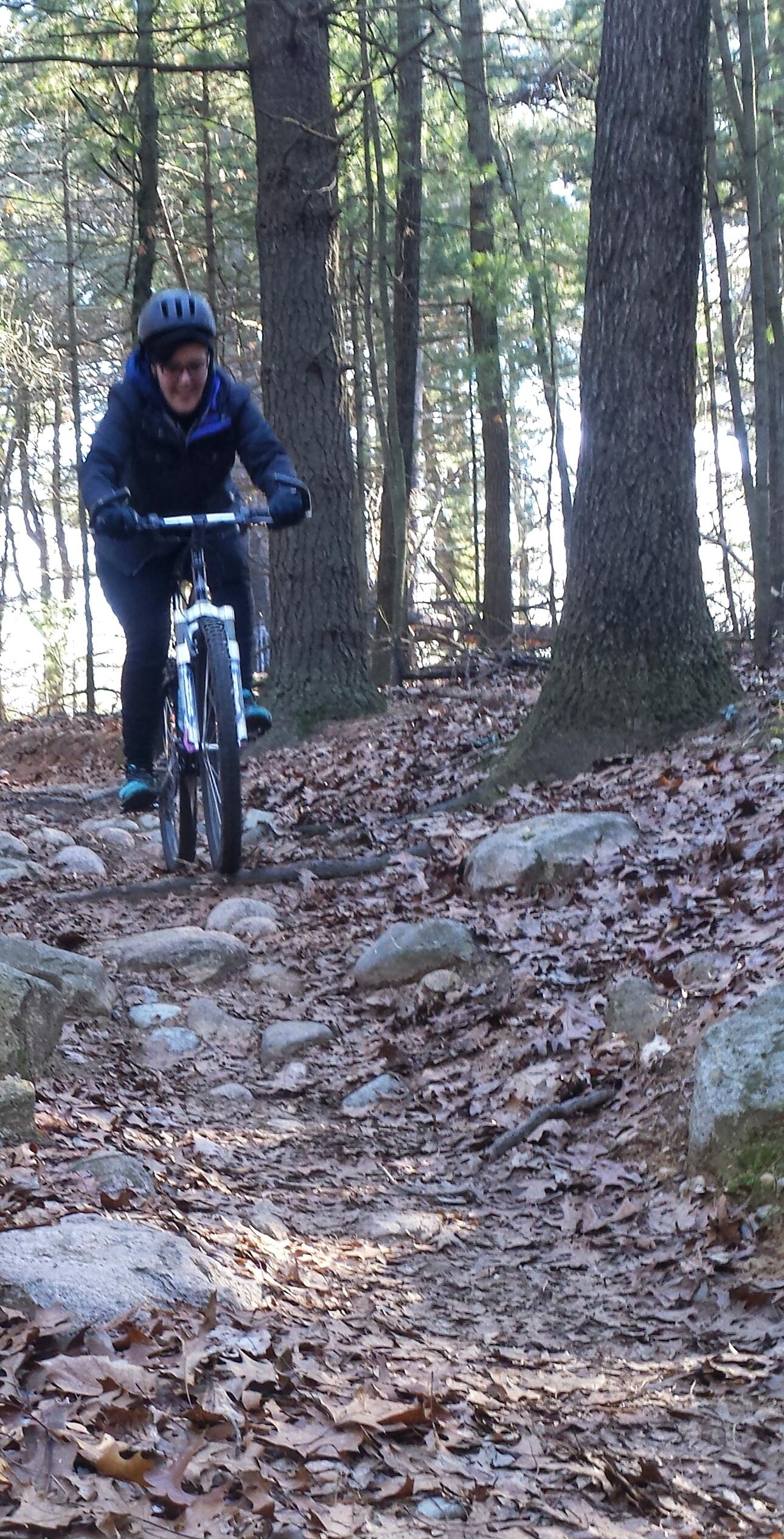 A person wearing a helmet and dark jacket rides a mountain bike along a rocky, leaf-covered trail in a wooded area. Tall trees surround the path, and the sunlight filters through the leaves. Yankee Springs mountain bike trail.