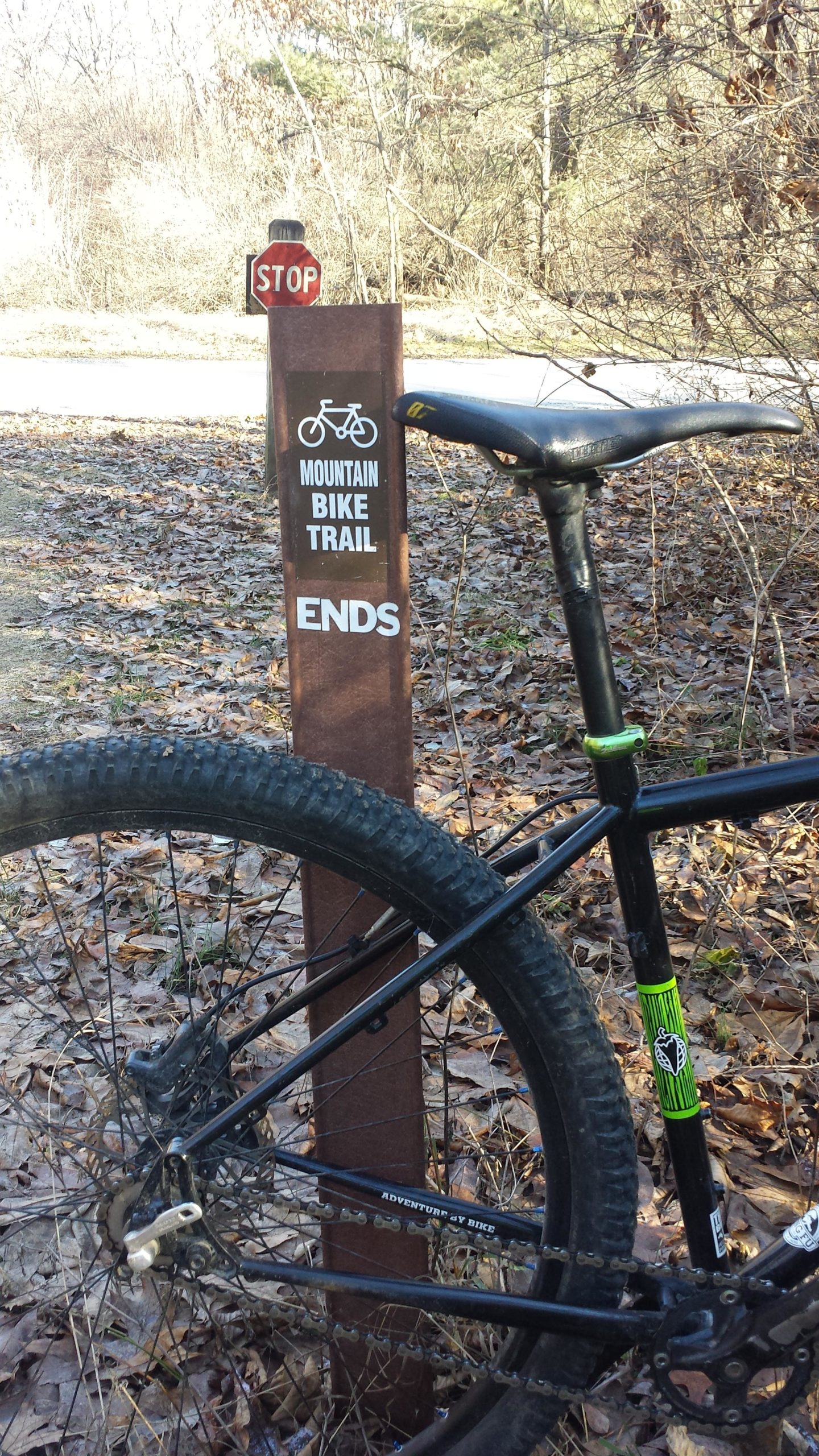 A mountain bike parked next to a trail marker indicating the end of a mountain bike trail. The sign reads "MOUNTAIN BIKE TRAIL ENDS" and is located beside a stop sign amidst a leaf-covered ground. Trees are visible in the background, indicating a natural outdoor setting. Yankee Springs mountain bike trail.