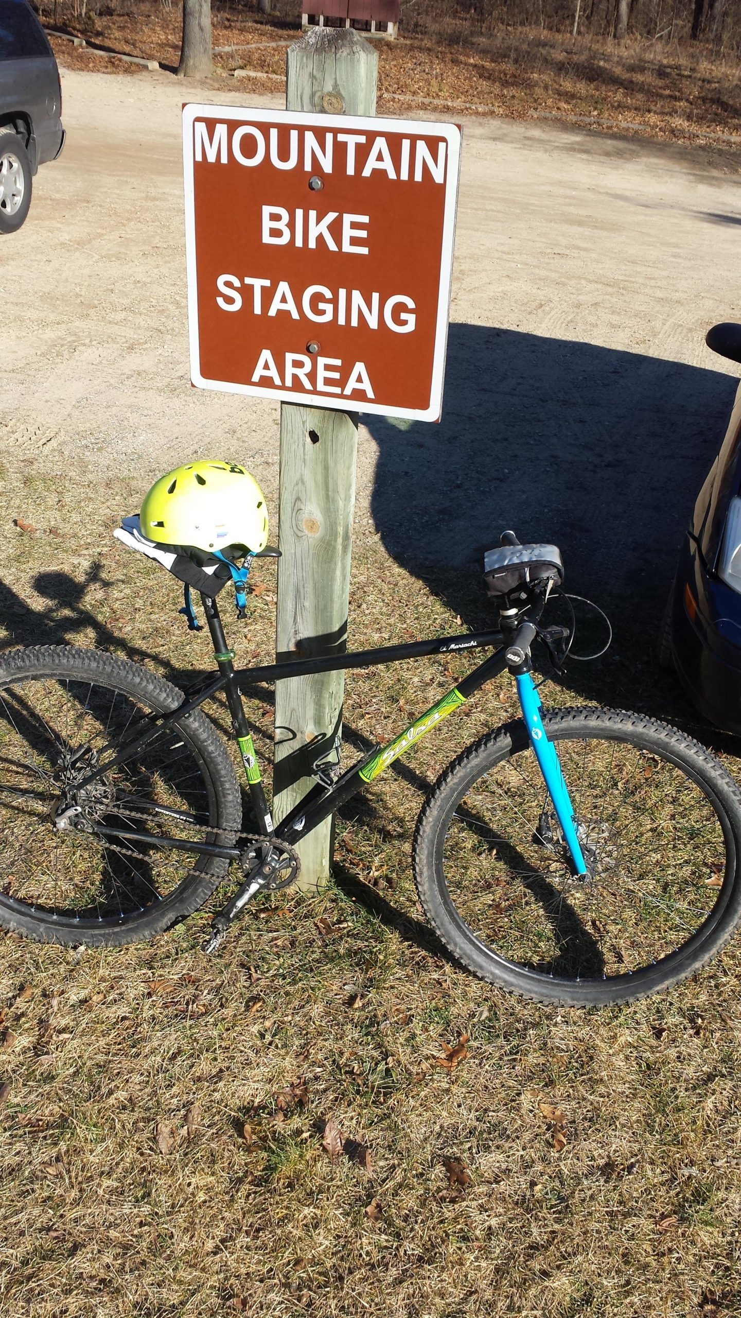 A mountain bike resting against a wooden post with a sign that reads "MOUNTAIN BIKE STAGING AREA." A bright yellow helmet is attached to the bike. The background features a dirt parking area and sparse trees, indicating an outdoor recreational setting. Yankee Springs mountain bike trail.